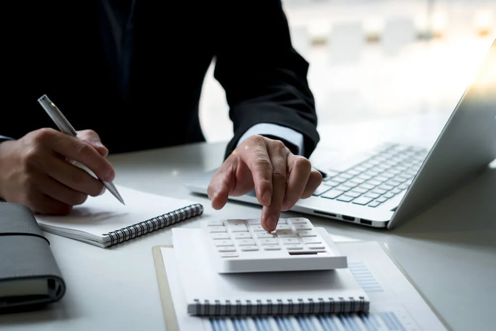 Barbat imbracat intr-un costum negru lucrand la un birou pe care se afla un laptop documente caiete si un calculator