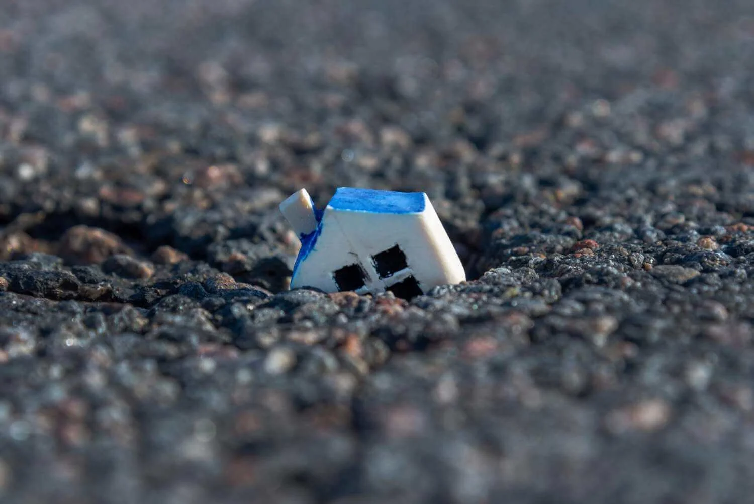 Casuta alba cu acoperis albastru partial ingropata in asfalt crapat