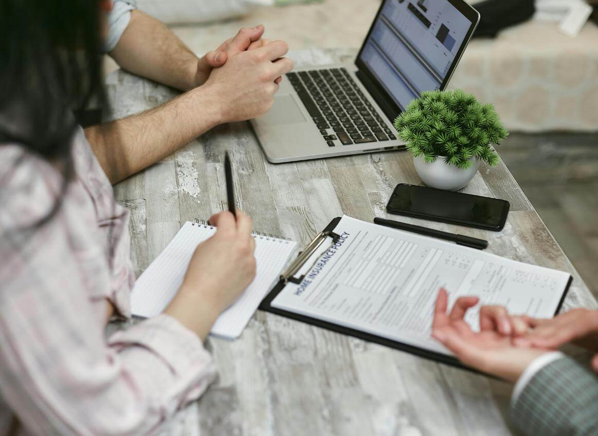 Trei persoane discuta la o masa cu laptop- formulare telefon oagenda si o planta decorativa