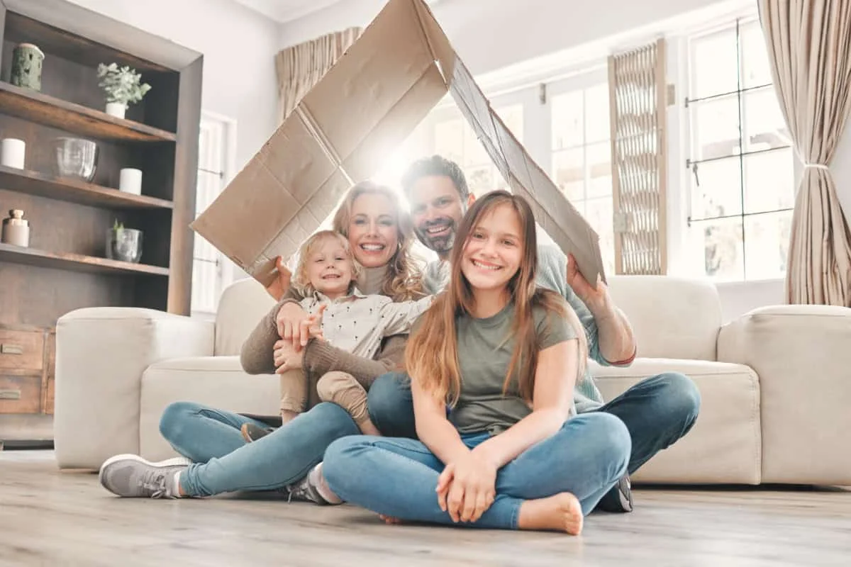 Familie fericita care sta pe podea in living tinand deasupra lor un carton in forma de acoperis