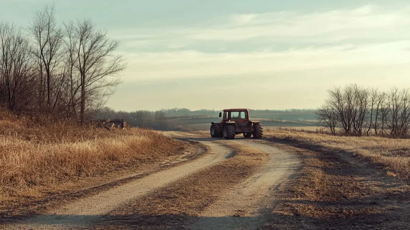 Tractor roșu pe un drum rural de pământ înconjurat de câmpuri și copaci desfrunziți