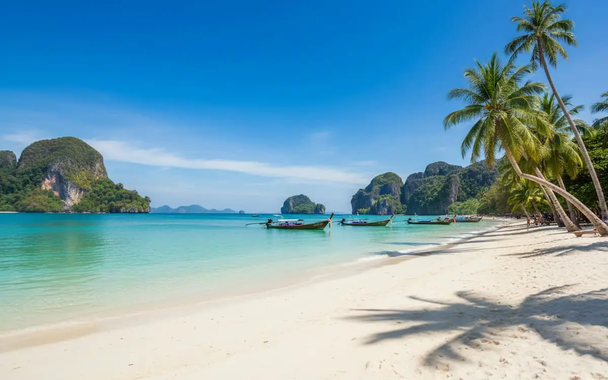 Plajă cu nisip alb, apă turcoaz, bărci tradiționale și palmieri în Thailanda